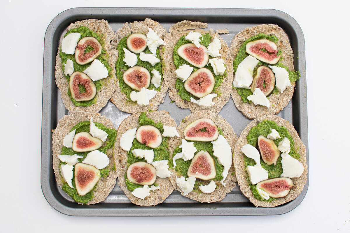 A baking tray with 4 halved pitta breads (8 halves) topped with green pesto, mozzarella and figs