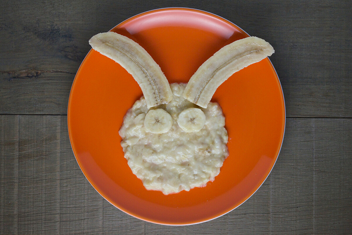 Pureed banana spread on plate in a circle to create face shape. Peeled and halved banana placed above face to create ears with banana rounds on face to create eyes