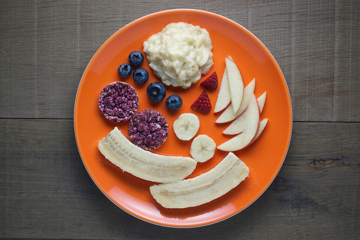 Apple cut into 6 thin slices, strawberry cut in half to create triangle pieces. 5 blueberries and rice cakes also on plate with banana puree and banana pieces