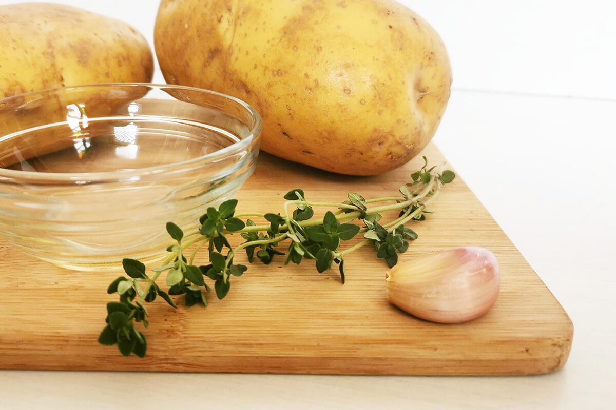 A chopping board with potatoes, garlic, rosemary and a little bowl of olive oil