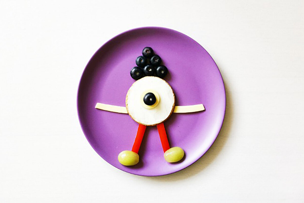 Oat cracker with two finger size slices of pepper creating legs, grapes creating feet and cheese slices for arms with a circular piece of cheese in the centre of oat cake. 6 blueberries are placed above it for hair and another in the centre of the oat cake and cheese, with a dollop of cream cheese or yoghurt, to create an eyeball