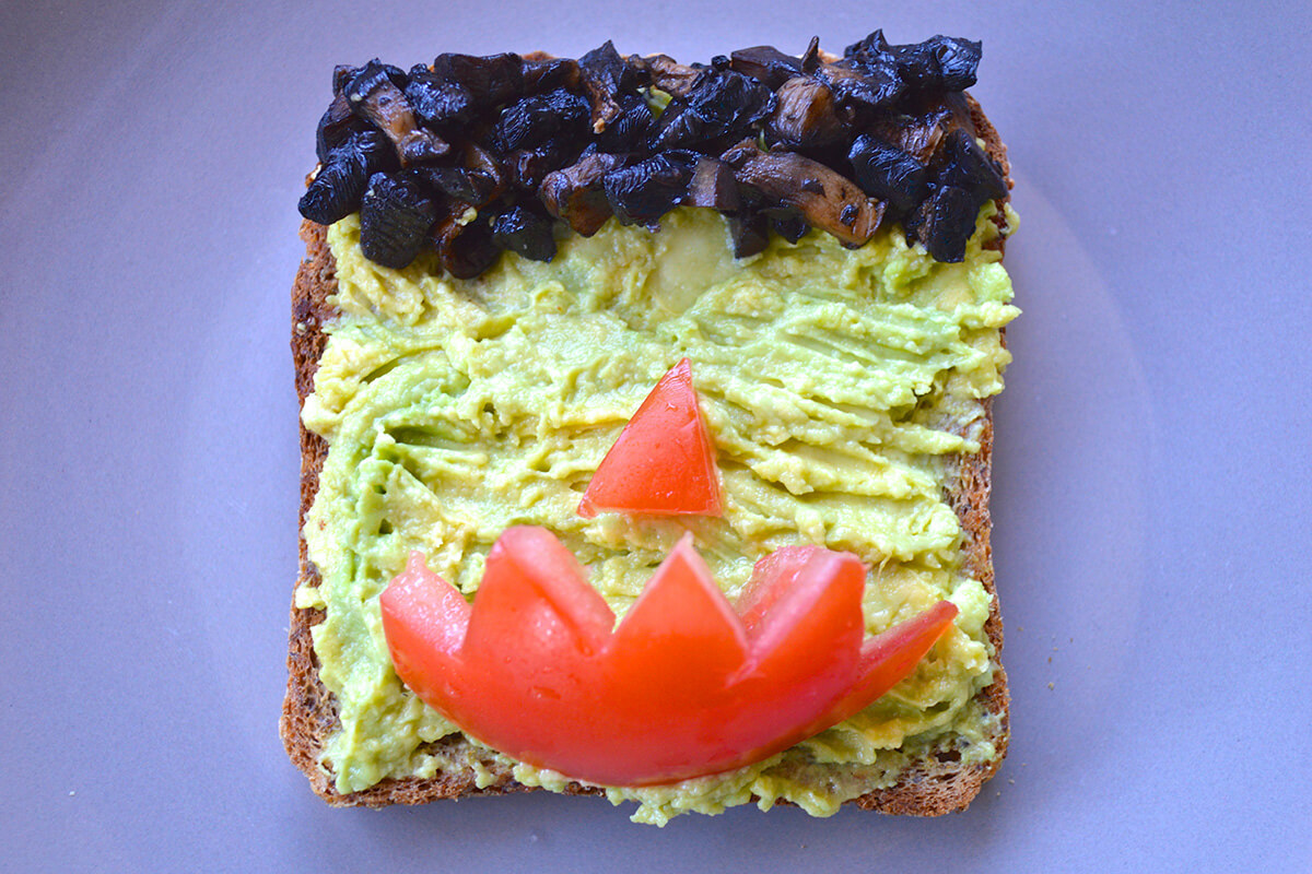 Toast topped with avocado, fried mushroom for hair and tomato for a nose and mouth