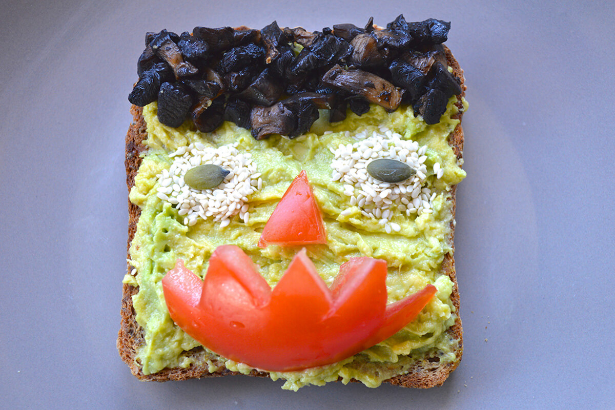 Toast topped with avocado, fried mushroom for hair, tomato for a nose and mouth and sesame seeds for eyes with pumpkin seed pupils