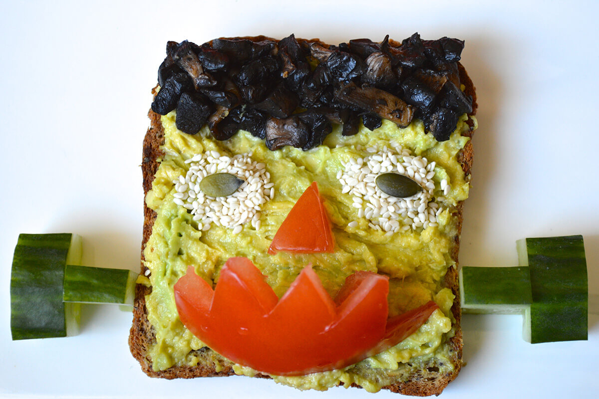 Halloween Frankenstein Toast with avocado skin, mushroom hair, sesame and pumpkin seed eyes, tomato nose and mouth and cucumber ears