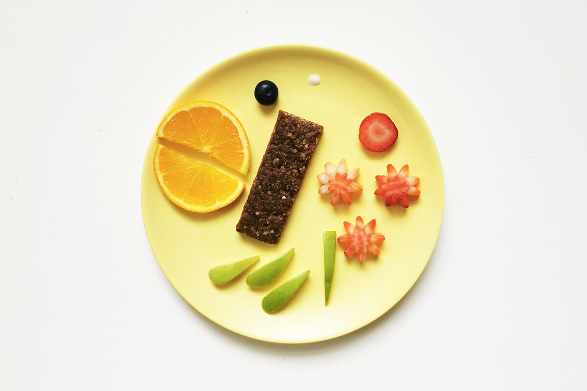 Sliced and halved orange slices arranged on a plate with 4 finger sized apple slices, a halved blueberry, sliced strawberry with 3 more strawberries slices cut into flower shapes