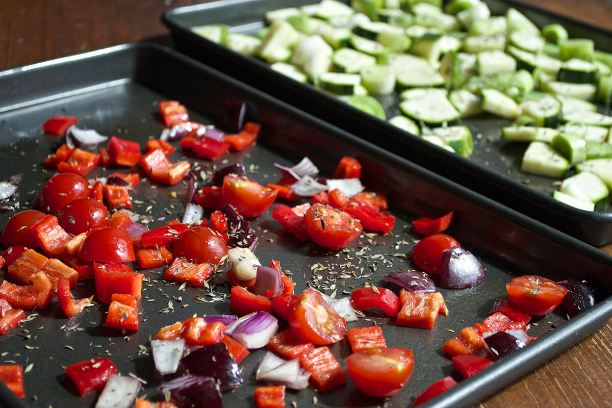 Two baking trays, one with leek and diced courgette, the other with chopped red onion, red pepper and cherry tomatoes