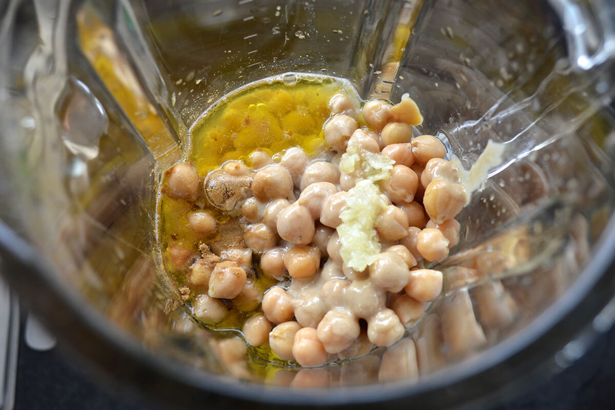 Chickpeas in a food processor with lemon juice, garlic cloves, cumin, tahini, water and extra virgin olive oil
