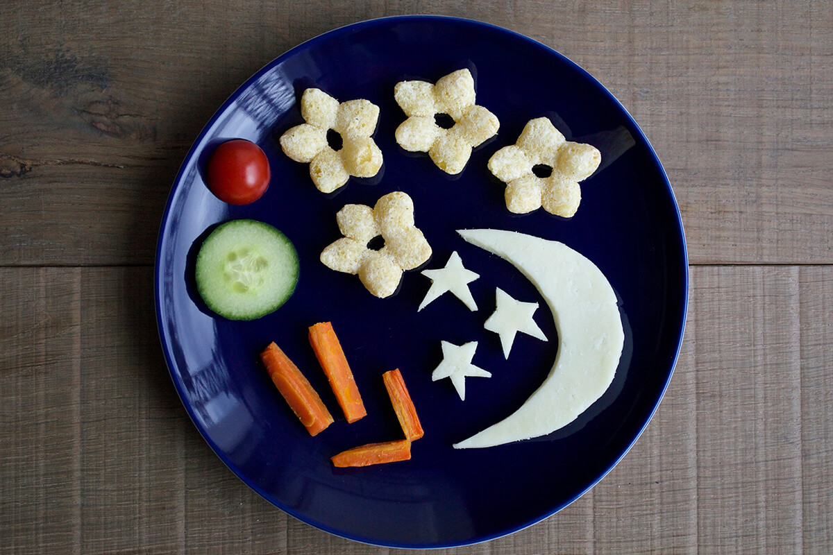Roasted peeled carrot batons arranged on plate with a cheese semicircle and stars, cucumber slice, cherry tomato and 4 organix melty cheese stars