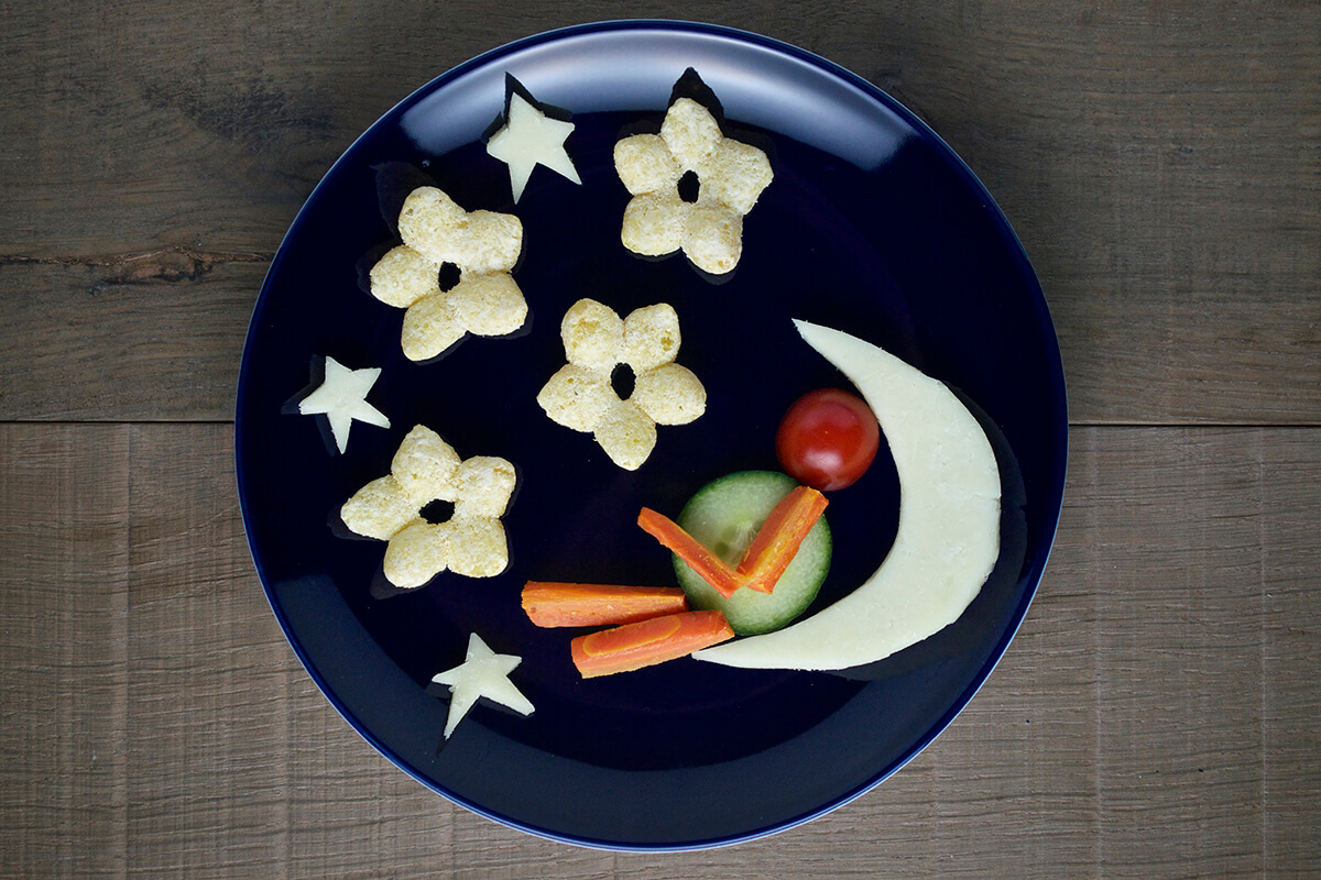 To complete the man on the moon fun plate, the cucumber slice is placed on the moon shape with the tomato on top to create a body and head. Two carrot sticks are placed under the cucumber slice to create legs and the other 2 carrot slices are on the cucumber, creating a bent arm shape
