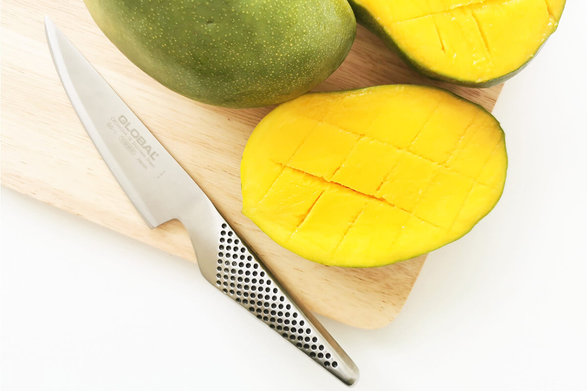 Whole mango on a chopping board next to a halved mango cut lengthways and crosswise without cutting through the skin