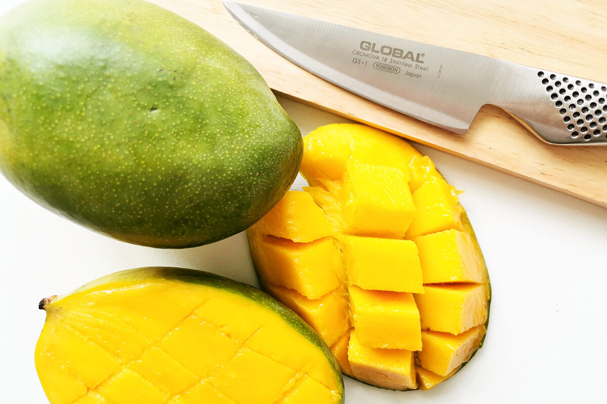 Whole mango on a chopping board next to a halved mango cut lengthways and crosswise without cutting through the skin and with the cubes pushed out of the mango