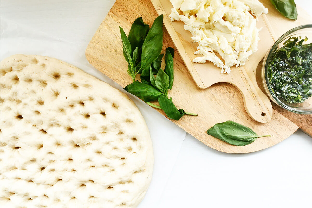 A wooden chopping board with mozzarella, fresh basil leaves and a small container of chopped basil mixed with olive oil next to dough with small holes on a baking sheet