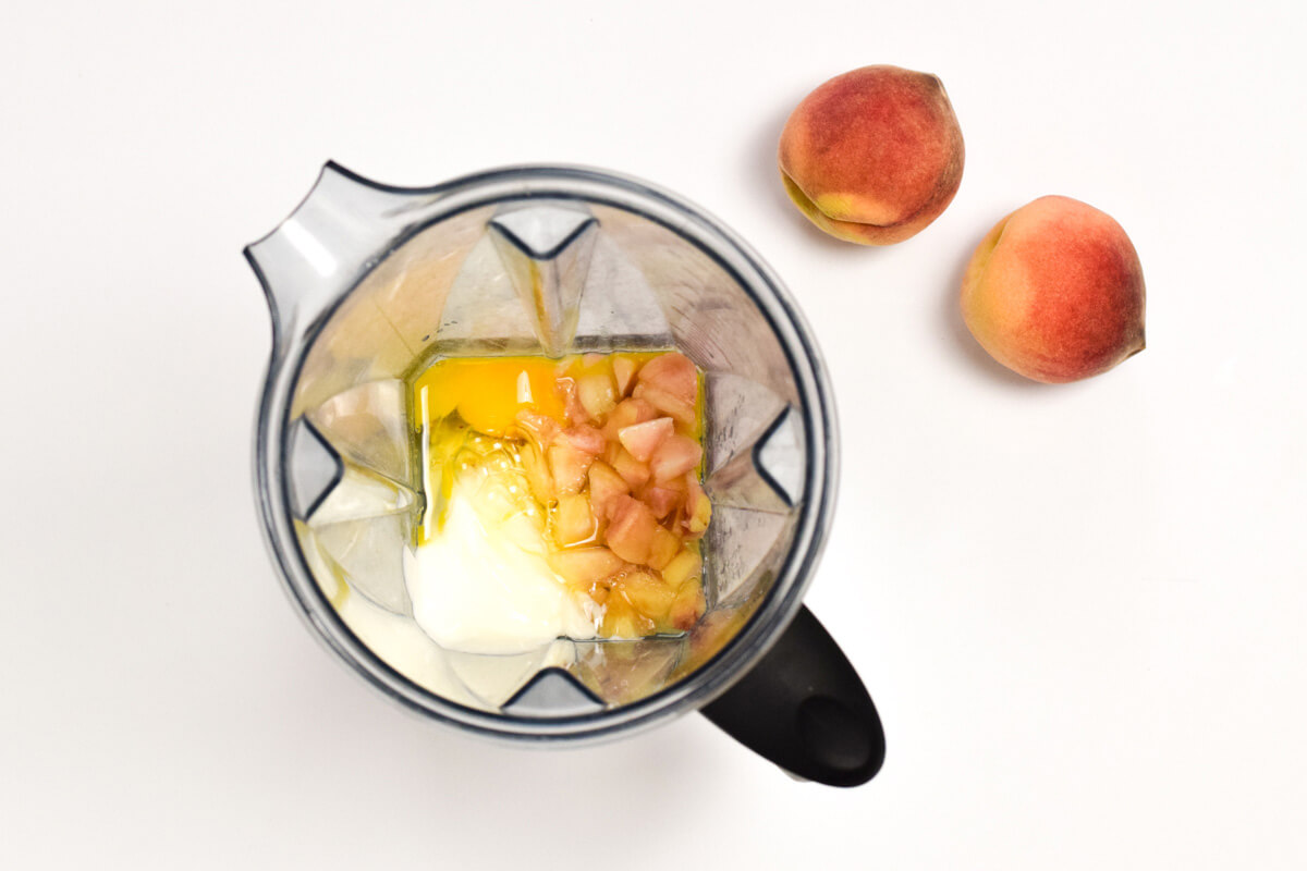 A food processor with oats, egg, diced peaches and yoghurt next to 2 whole peaches