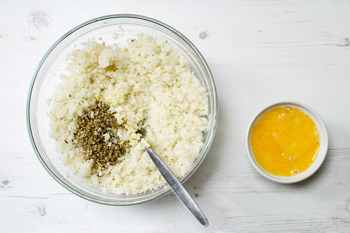 Cauliflower rice in a glass bowl with oregano and crushed garlic, next to a small bowl of beaten eggs