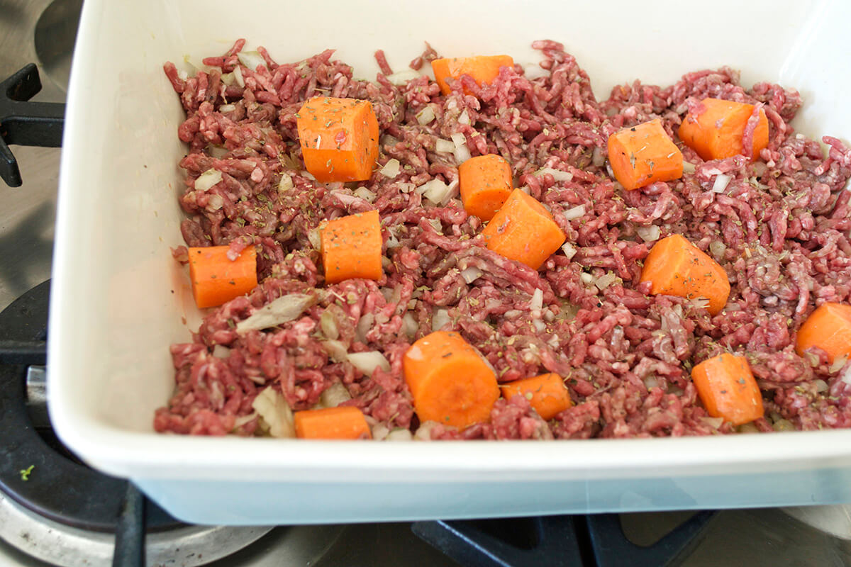 A casserole dish with raw mince, finely chopped onion, carrots, pepper and herbs