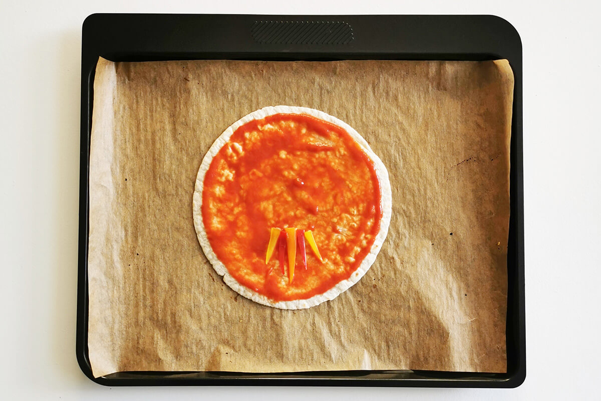 A lined baking tray with a tortilla wrap topped with pizza sauce and peppers at the bottom of the wrap to create flames