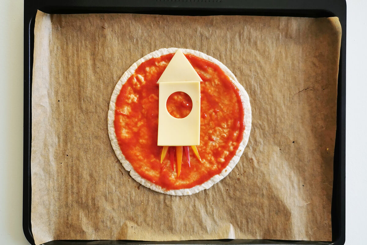 A lined baking tray with a tortilla wrap topped with pizza sauce and peppers at the bottom of the wrap to create flames and two cheese slices shaped and arranged to create a rocket