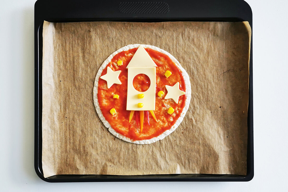 A lined baking tray with a tortilla wrap topped with pizza sauce and peppers at the bottom of the wrap to create flames and two cheese slices shaped and arranged to create a rocket, cheese slices shaped like stars and a sprinkle of sweetcorn