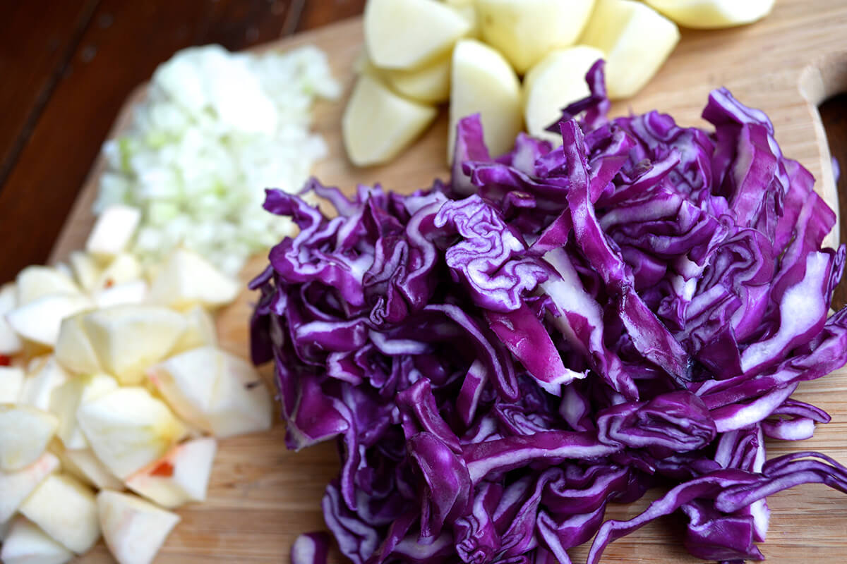A chopping board with chopped cabbage, potatoes, apple and onion