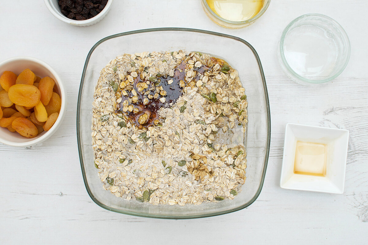 A bowl of rolled oats, oat flour, cinnamon, ginger, sunflower seeds and pumpkin seeds with maple syrup, coconut oil and vanilla extract next to a bowl of apricots and raisins and some small, empty bowls