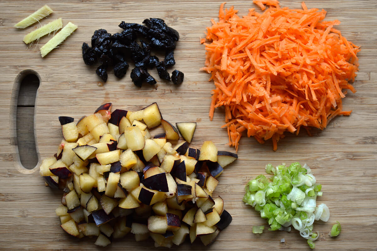 A chopping board with sliced ginger, chopped prunes, grated carrot, chopped spring onion and plums