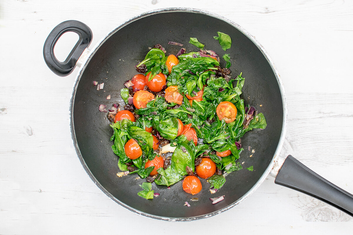 A wok with olive oil, chopped onion and crushed garlic, cherry tomatoes, raisins and spinach