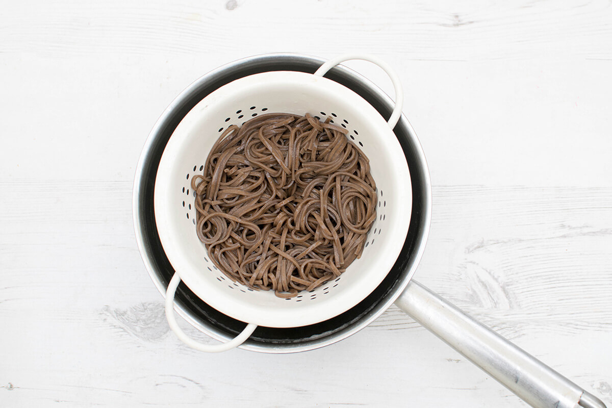 Soba noodles being drained in a colander over a saucepan
