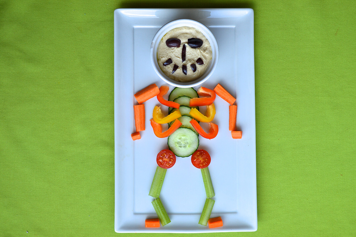 Spooky Skeleton Platter with vegetable body and a bowl of hummus with olives as a face