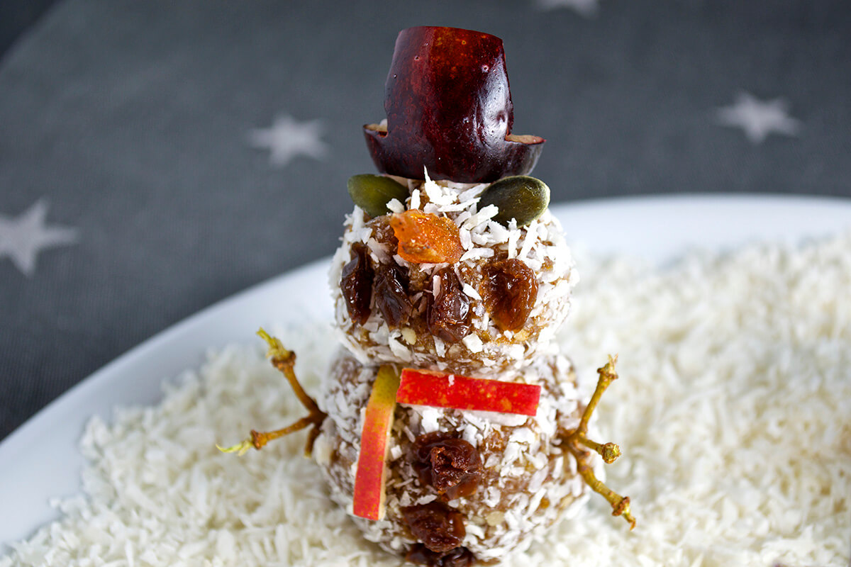 Assembled Sweet Treat Snowman, made of the nutty date mix rolled into balls on a cocktail stick, a grape for a hat, pumpkin seed eyes, dried apricot for a nose, a slither of apple for a scarf and halved raisins for a smile and buttons with grape stems for arms, sat on desiccated coconut 'snow'