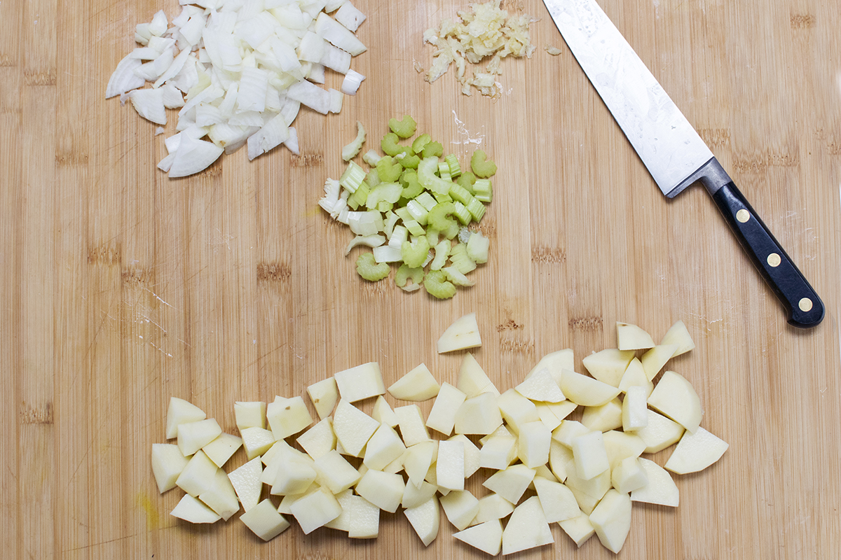 A chopping board with peeled and diced onion, crushed garlic cloves, sliced celery stalk and peeled and diced potatoes