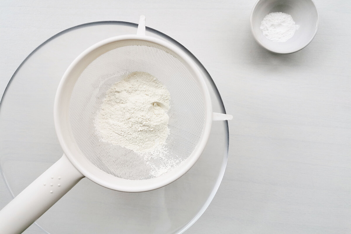 Flour being sieved into a glass bowl with a small bowl of baking powder next to it