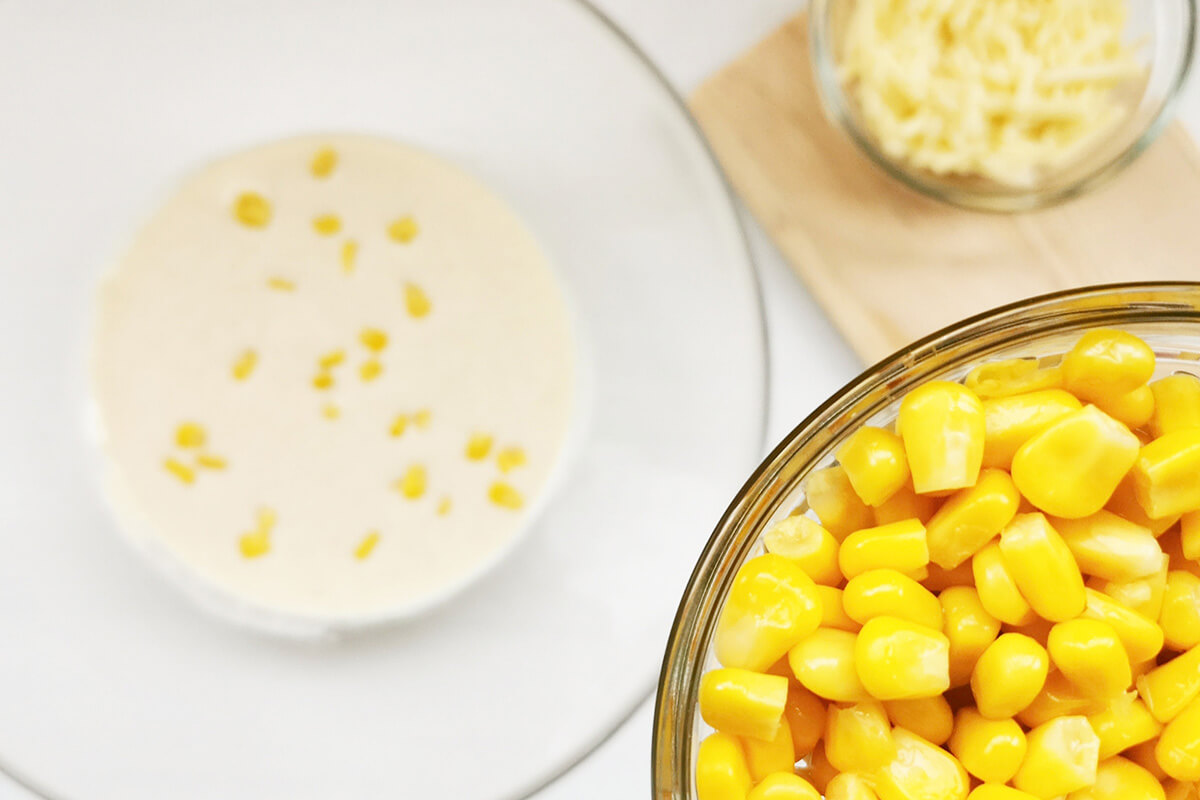A glass bowl of egg and milk next to a bowl of sweetcorn and a small bowl of grated cheese on a chopping board