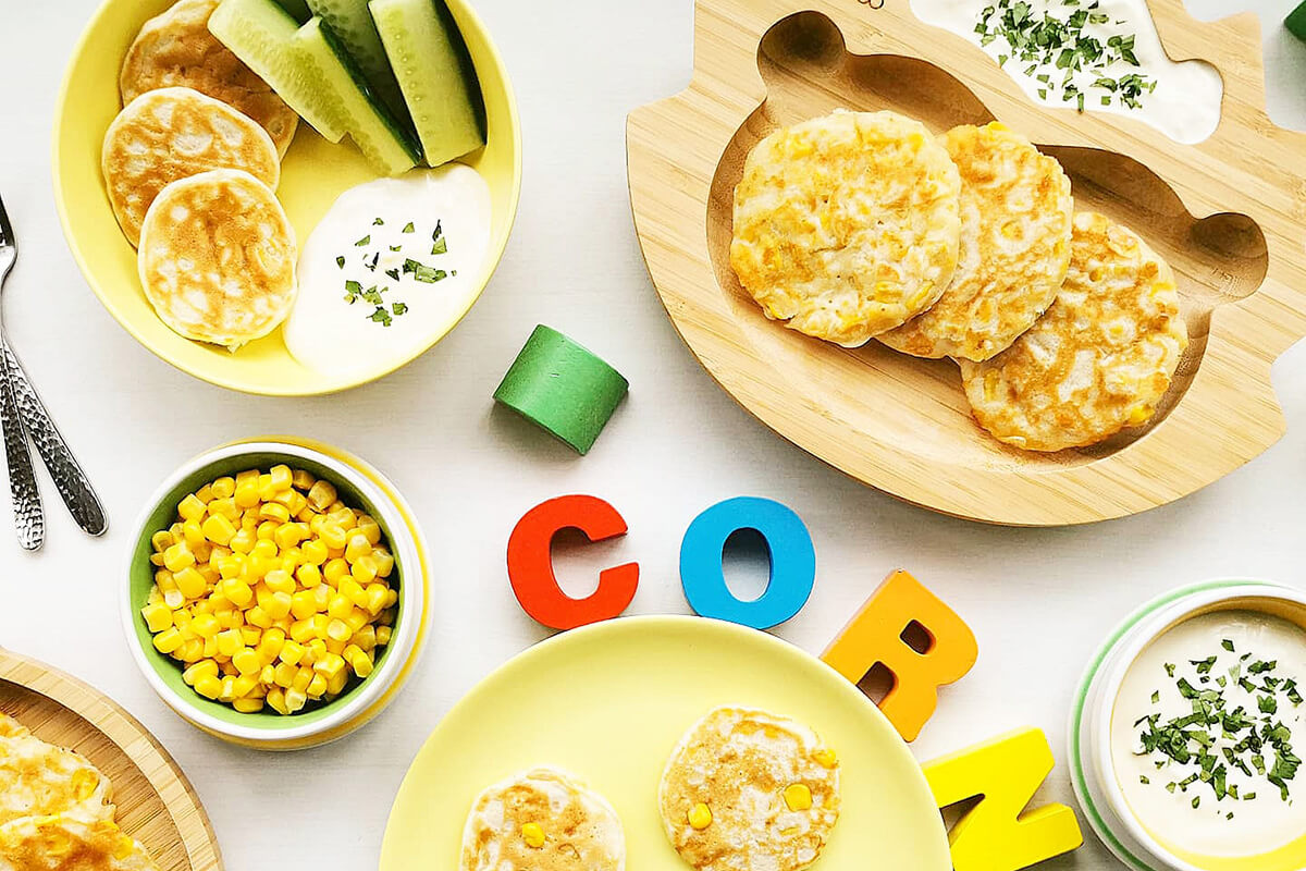 4 servings of sweetcorn fritters served with a yoghurt dip and next to a bowl of sweetcorn