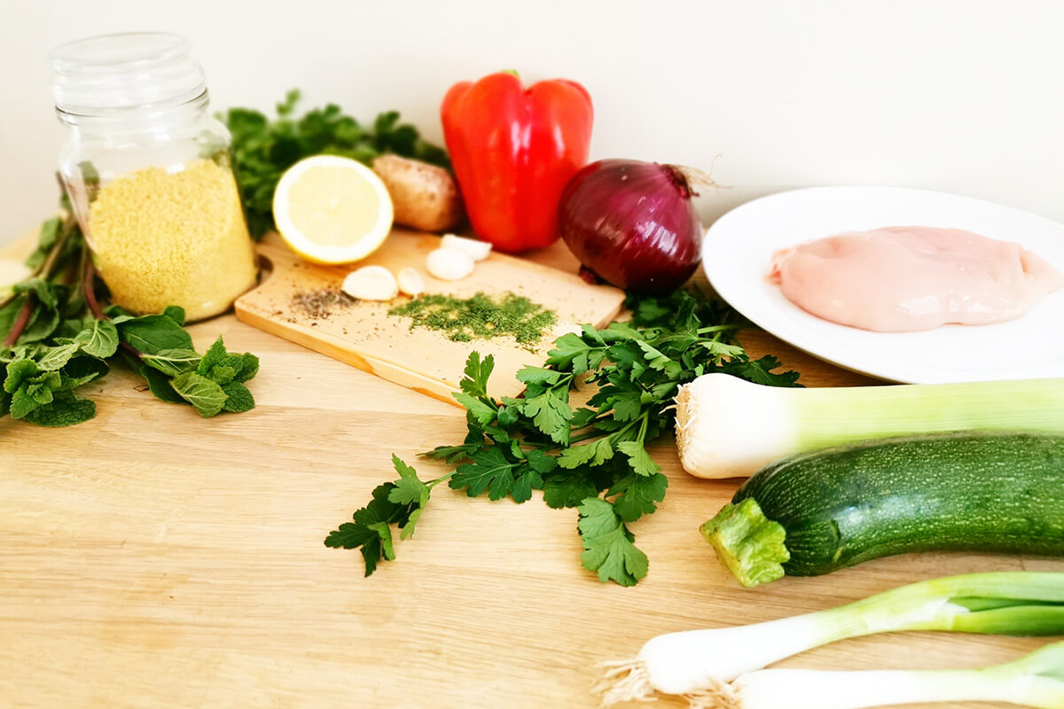 A chopping board with a container of uncooked couscous, raw chicken on a plate, courgette, leek, spring onion, red ocion, red pepper, have a lemon, some garlic cloves, coriander and some mint leaves