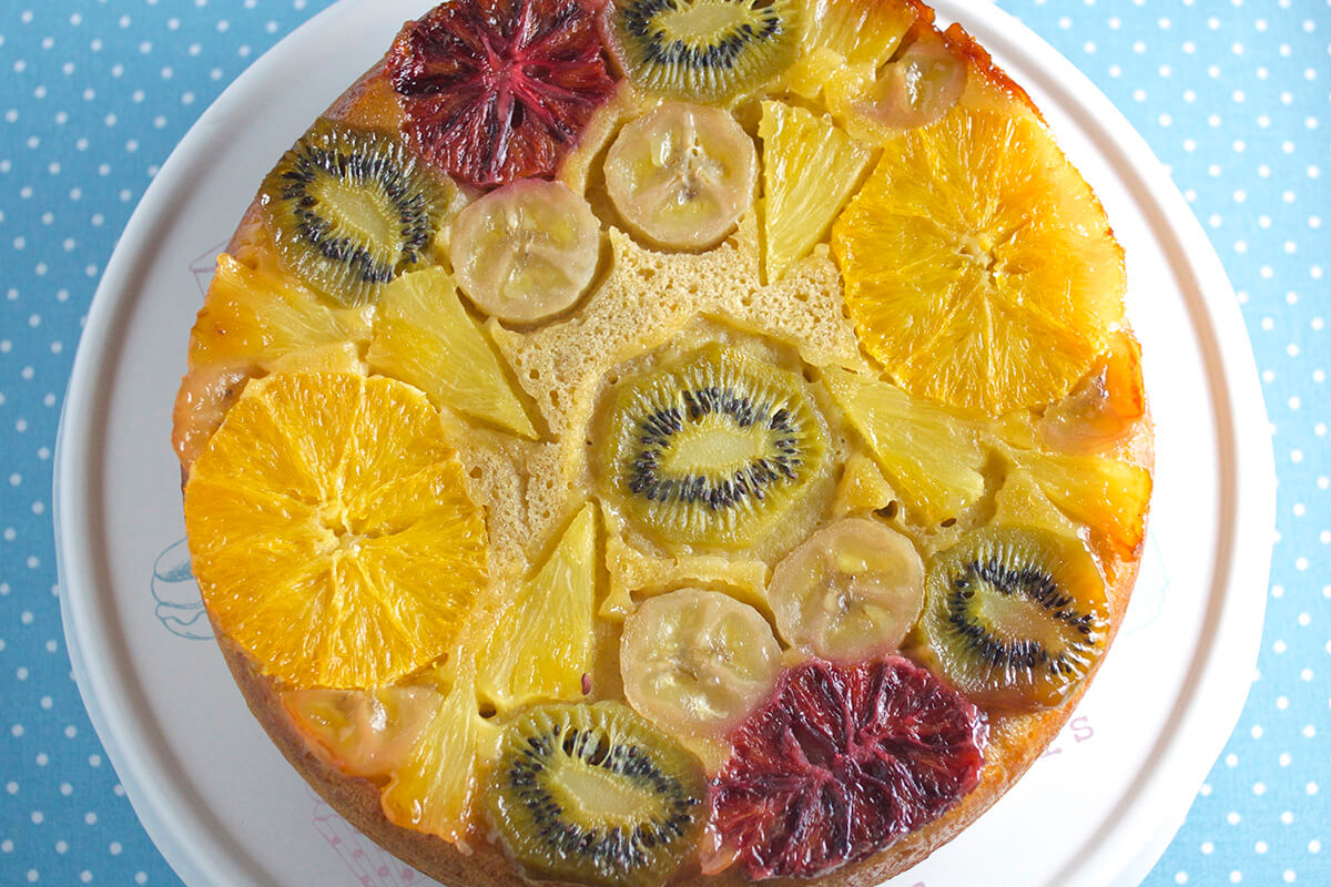 Tropical upside down cake on a cake stand. The bottom of the cake has kiwi, orange, blood orange, banana and pineapple chunks