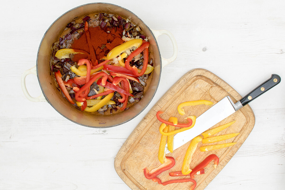 A saucepan with chopped onion, red and yellow peppers, crushed garlic, ground cumin and smoked paprika next to a chopping board with sliced red and yellow pepper