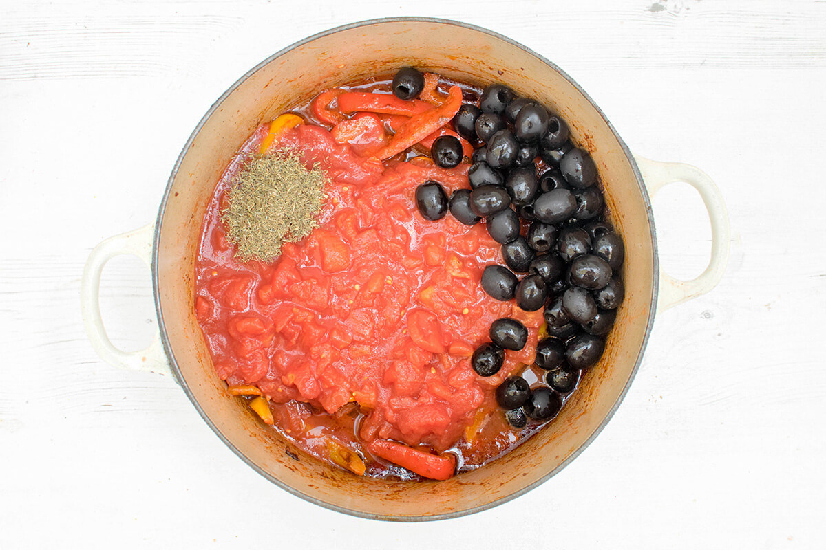 A saucepan with chopped onion, red and yellow peppers, crushed garlic, ground cumin and smoked paprika, chopped tomatoes, thyme, chilli powder and olives