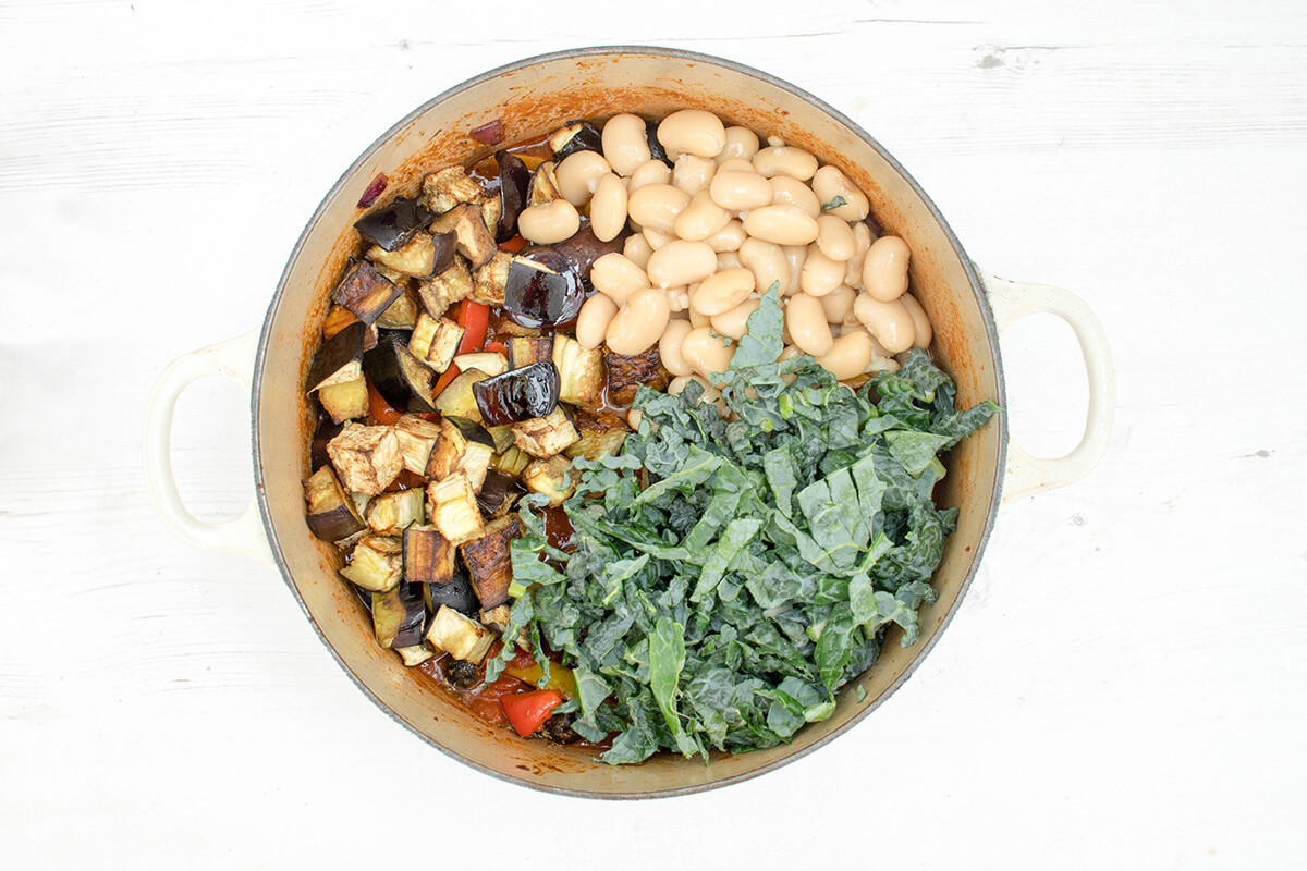 A saucepan with chopped onion, red and yellow peppers, crushed garlic, ground cumin and smoked paprika, chopped tomatoes, thyme, chilli powder and olives, butter beans, cider vinegar and chopped kale