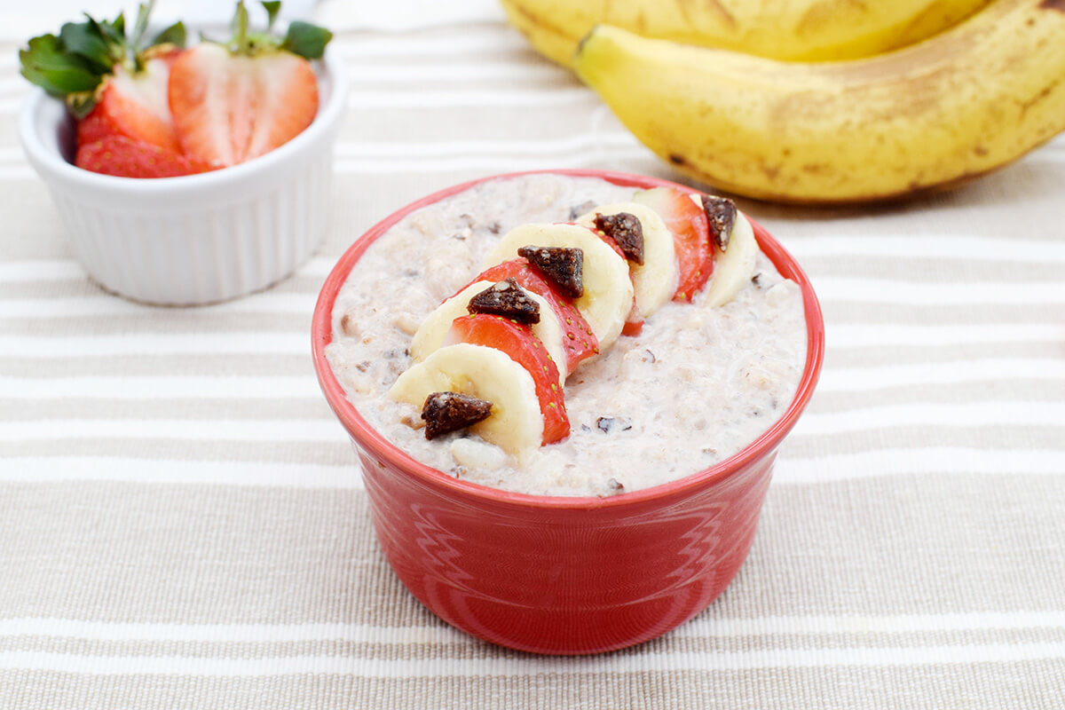 A bowl of rice pudding topped with sliced strawberries and bananas and Organix Gummies, next to a ramekin of sliced strawberries and some bananas