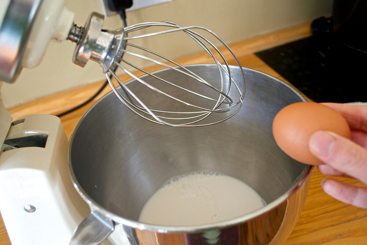 An egg being broken into a bowl of milk beneath and electric whisk