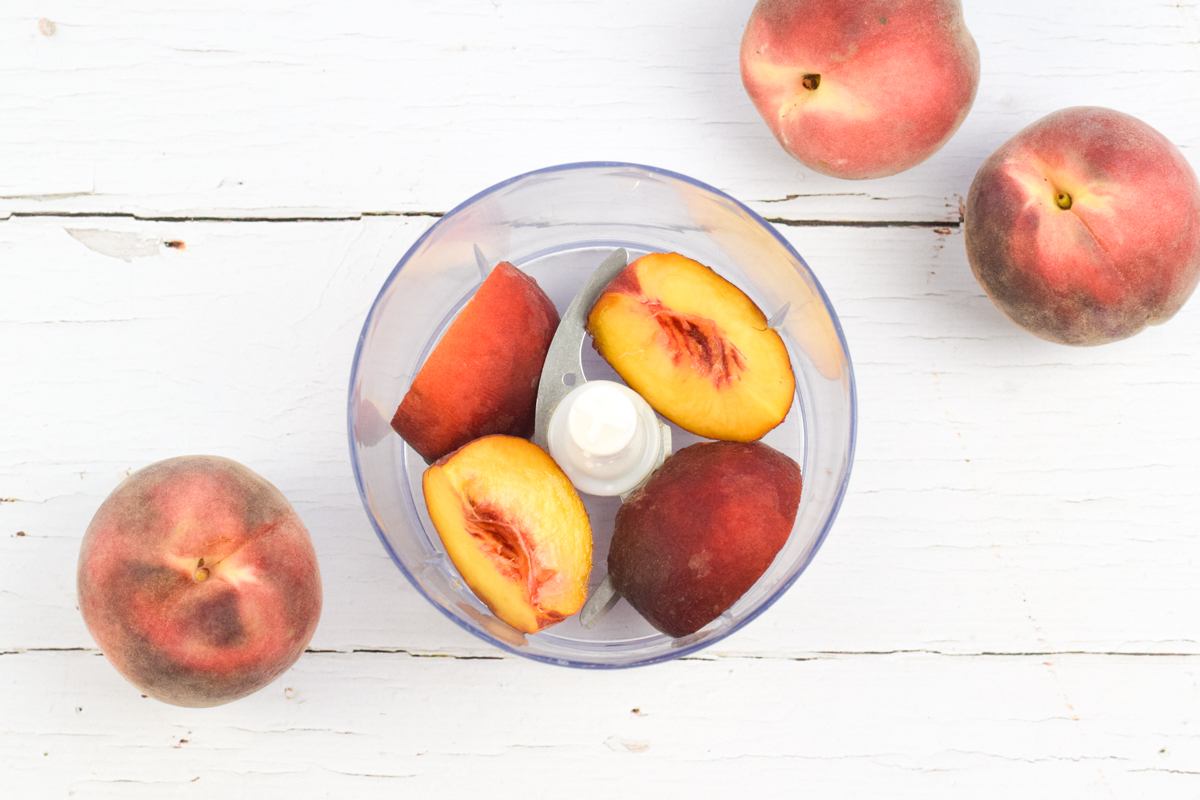 A food processor with a quartered peach, next to 3 peaches