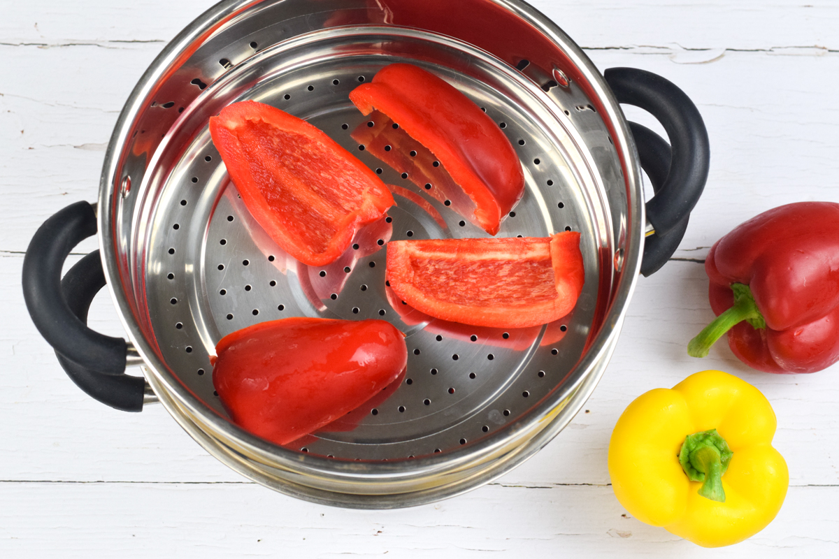 A quartered red pepper in a steamer, next to a whole yellow and red pepper