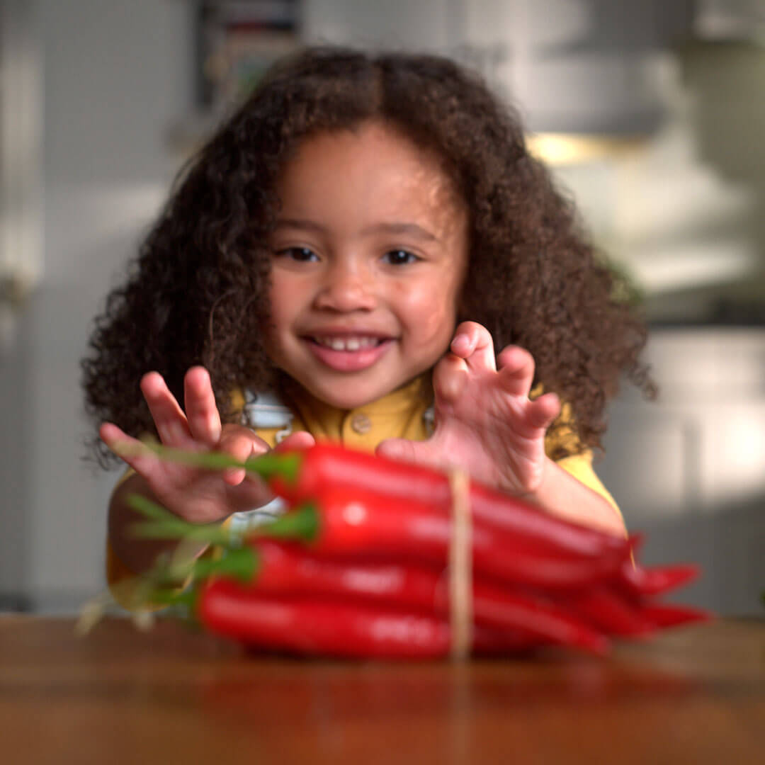 A toddler smiling in front of a table with some red peppers